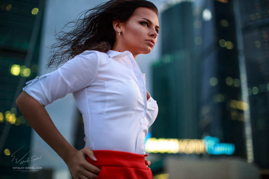 Evening city lights and beautiful brunette in white shirt and red skirt moving forward from left to right. Closeup half body portrait.